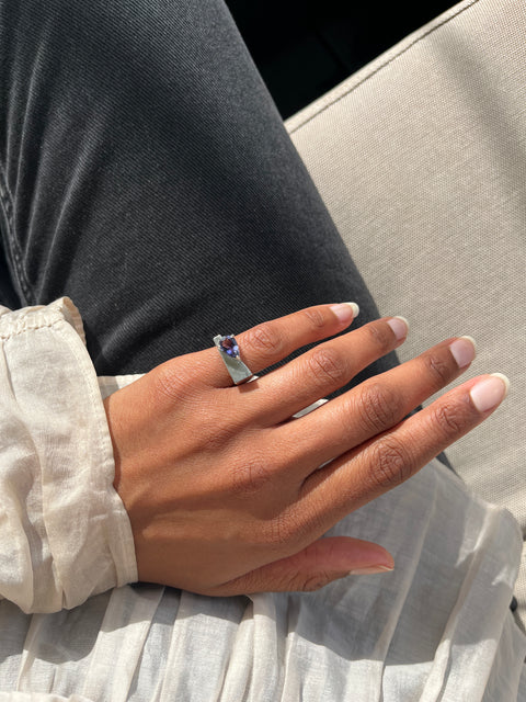 Hand wearing a ring with a colorful gemstone, resting on a textured surface.