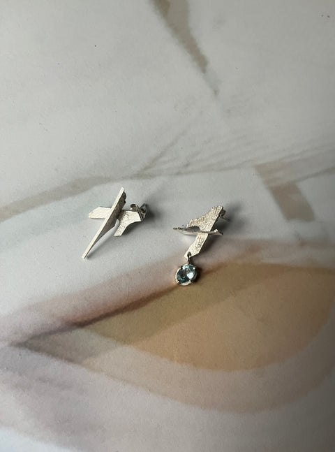 A pair of silver 'Aero' earrings with an asymmetrical design and one side featuring an aquamarine gemstone, displayed on a piece of fabric.