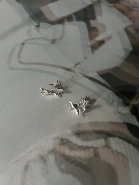 A pair of silver 'AERO MINI' earrings with an asymmetrical design, displayed on a textured surface.