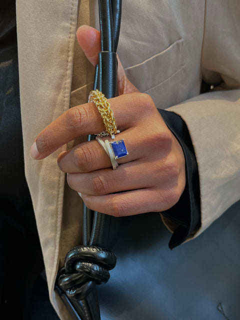 Hand wearing a gold and blue ring on a leather strap, with a person in a beige coat.